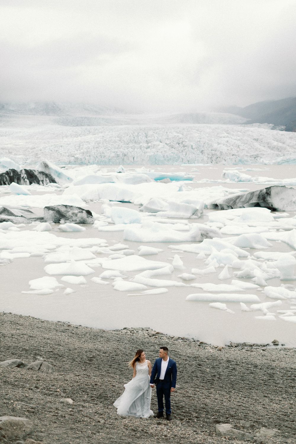 Bride and groom standing hand in hand on a dark shore with a breathtaking glacial lagoon and icebergs behind them.