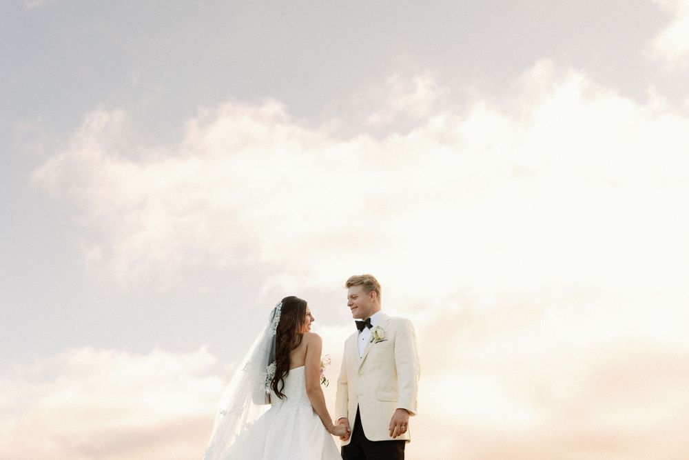 Bride in a long veil and white gown holds hands with groom in a white dinner jacket and bow tie against a soft pastel sky.