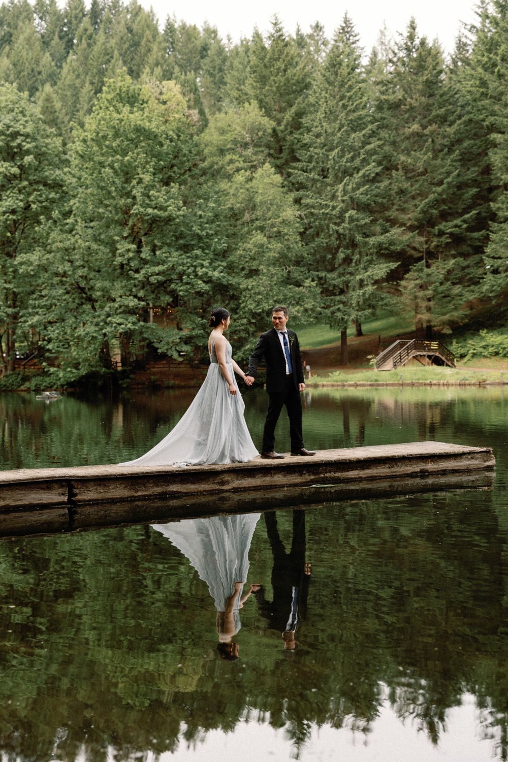 Bride and groom hold hands on a wooden dock reflected in a serene forest lake.