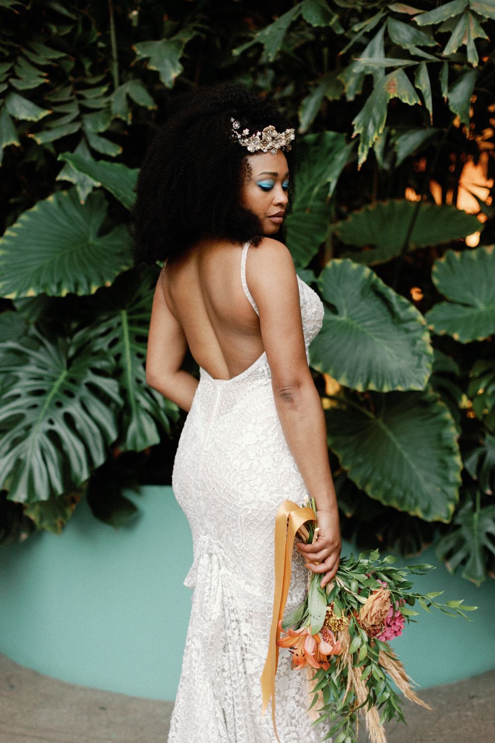 Black bride in backless lace gown wearing a crown holding a tropical bouquet outdoors.