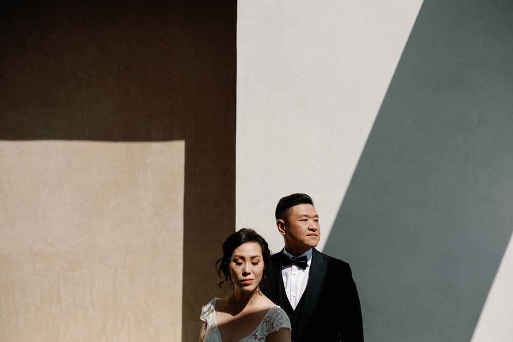 Elegant couple in formal wedding attire posing against geometric shadows on a minimalist wall.