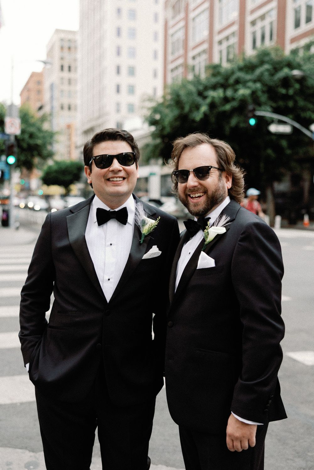 Two groomsmen in black tuxedos and sunglasses smiling on a city street crossing.
