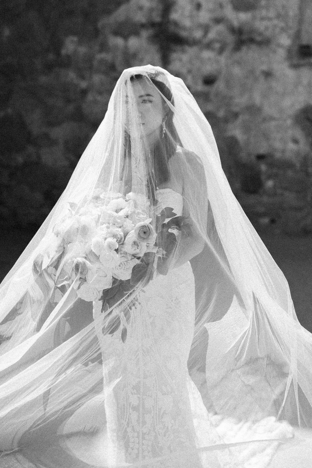 Black and white portrait of bride in lace gown holding bouquet beneath flowing wedding veil.