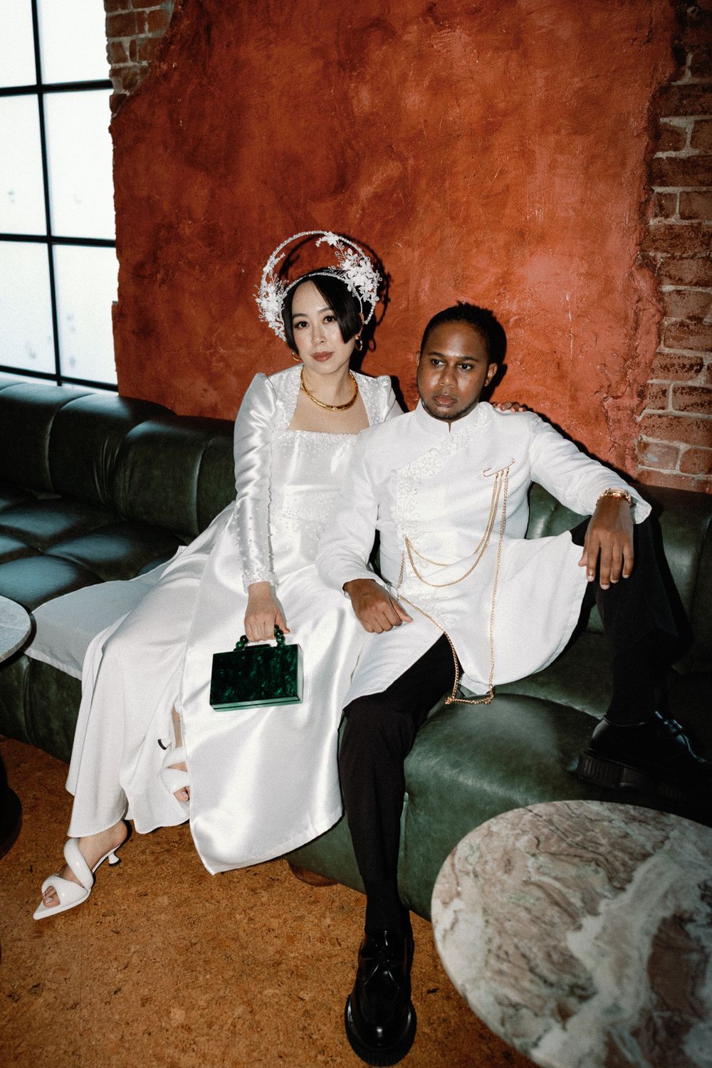 A bride in a traditional Vietnamese ao dai and groom in white sit together on a green leather sofa.
