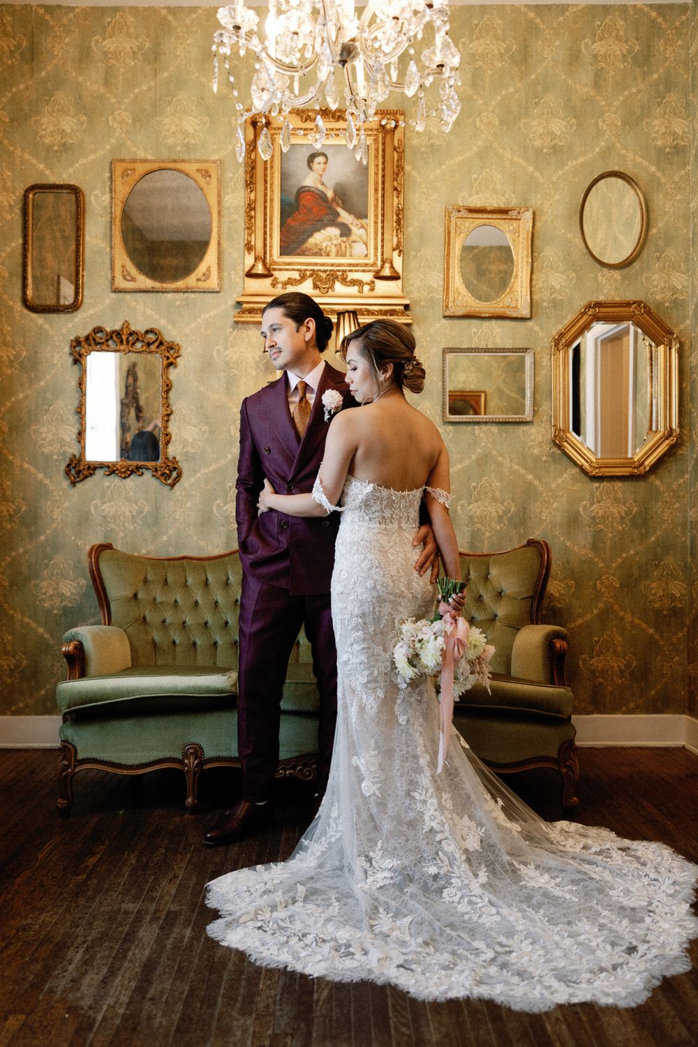 Bride in lace gown and groom in purple suit pose in vintage room with gold mirrors.