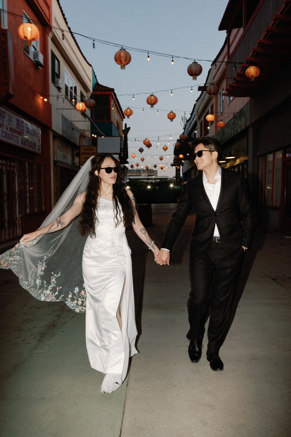 Bride in a white gown with a long lace veil and groom in a black suit walk hand in hand under hanging red lanterns on a city street at dusk.
