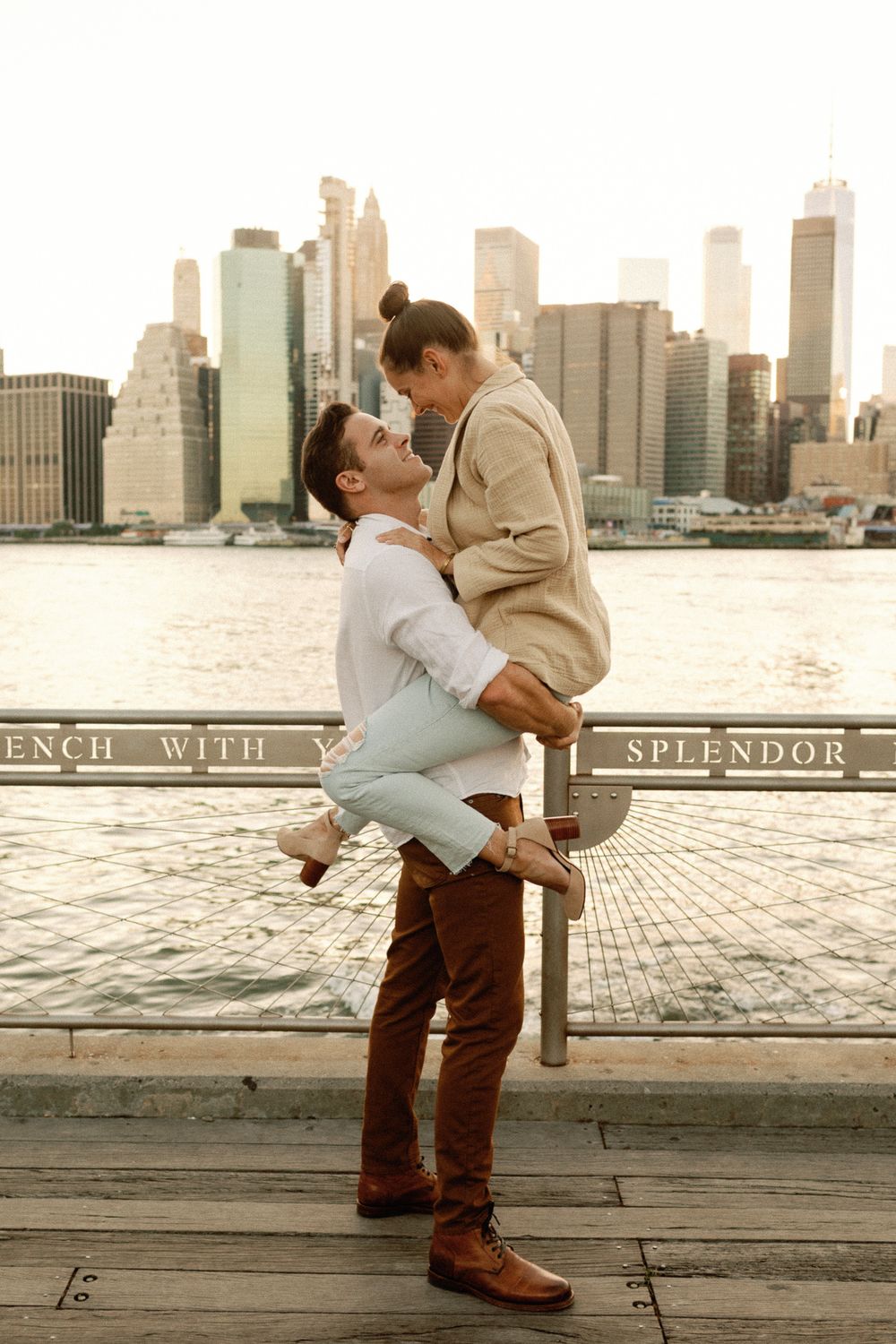 Man lifting woman in his arms on a waterfront boardwalk with NYC skyline at sunset.