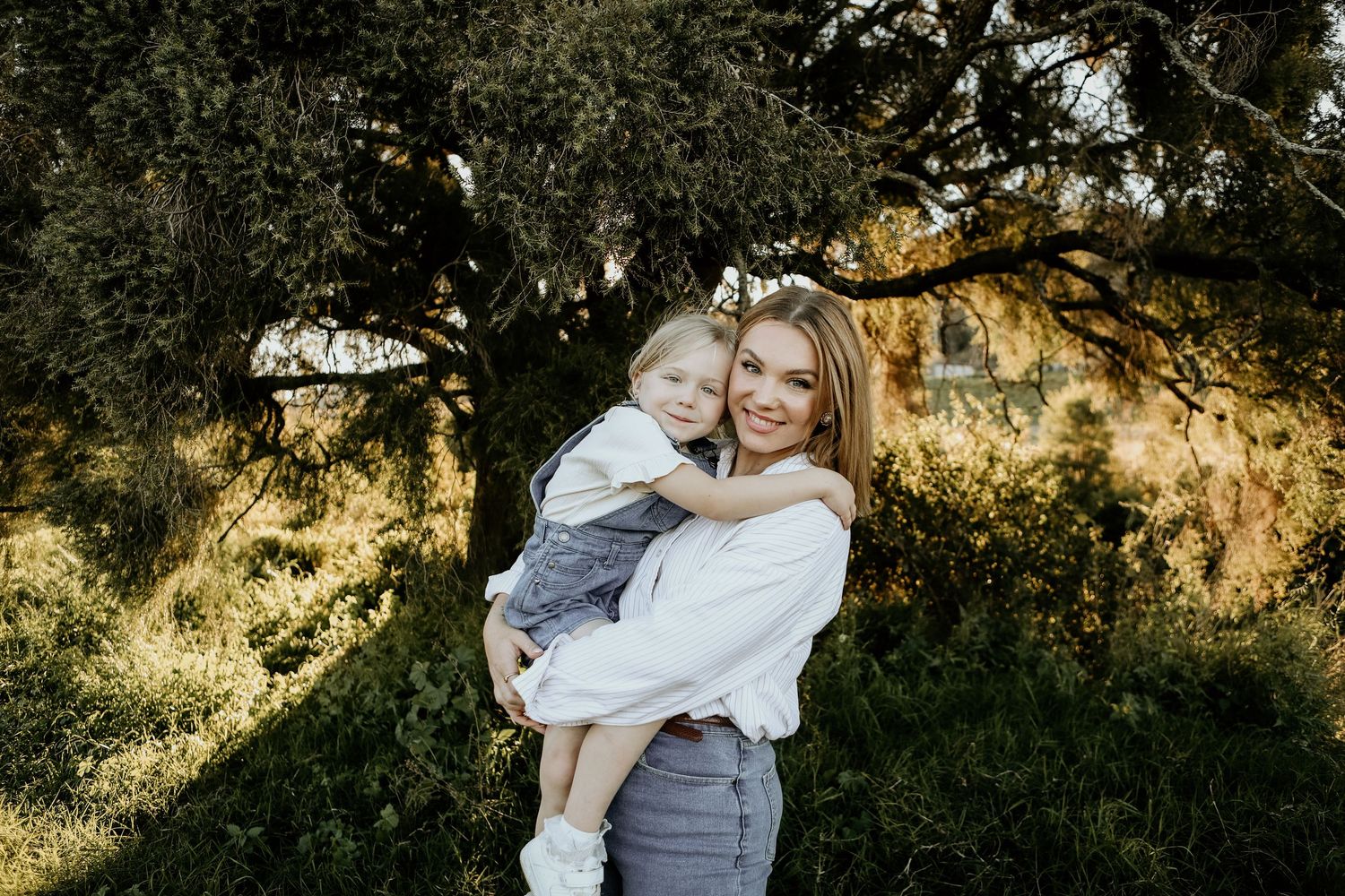 A parent and child share a joyful embrace outdoors against a sunlit natural backdrop with trees and foliage.