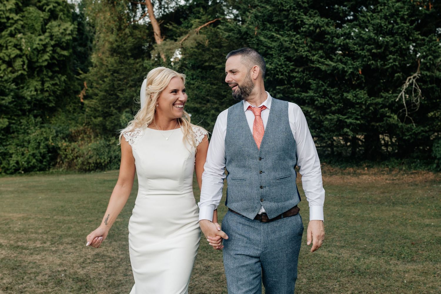 A couple in wedding attire walks together outdoors on their special day surrounded by lush green trees and foliage.