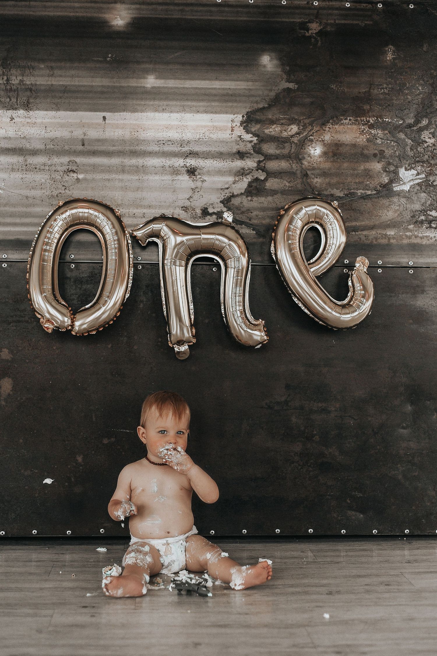Baby wearing diaper plays with silver letter balloons on dark wooden floor during cake smash photoshoot.