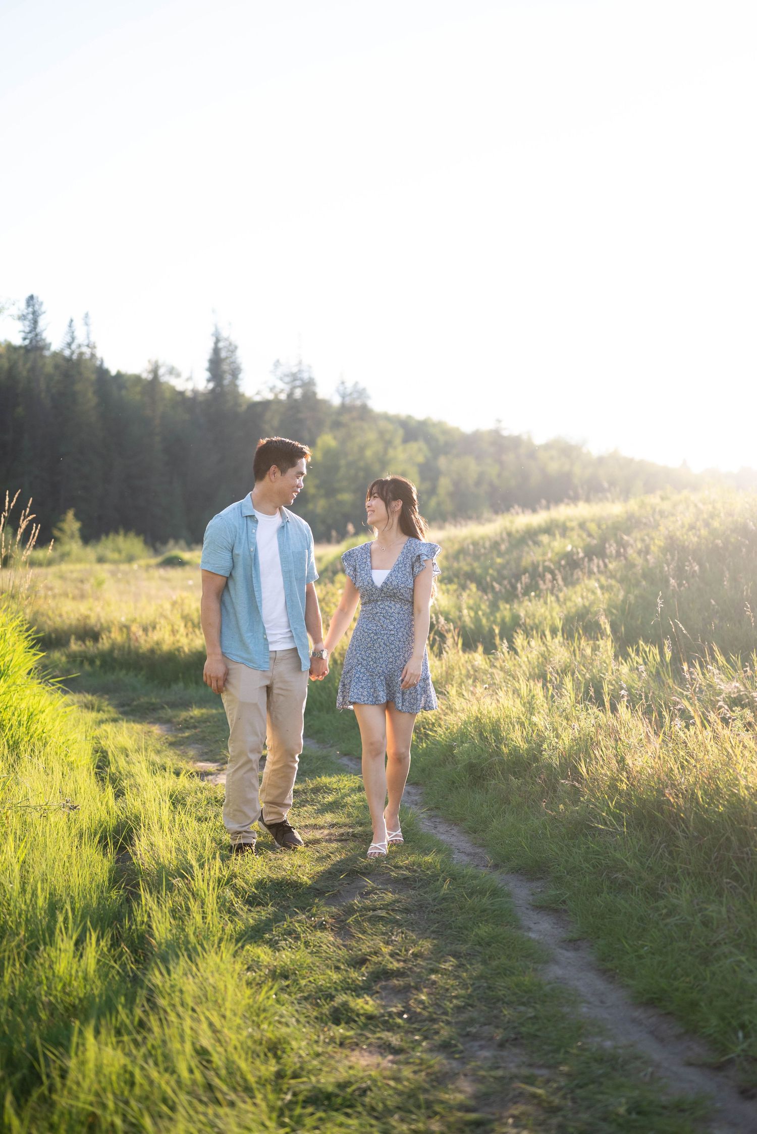 Two people stroll along a scenic mountain trail during sunset with tall grass and trees in the background.