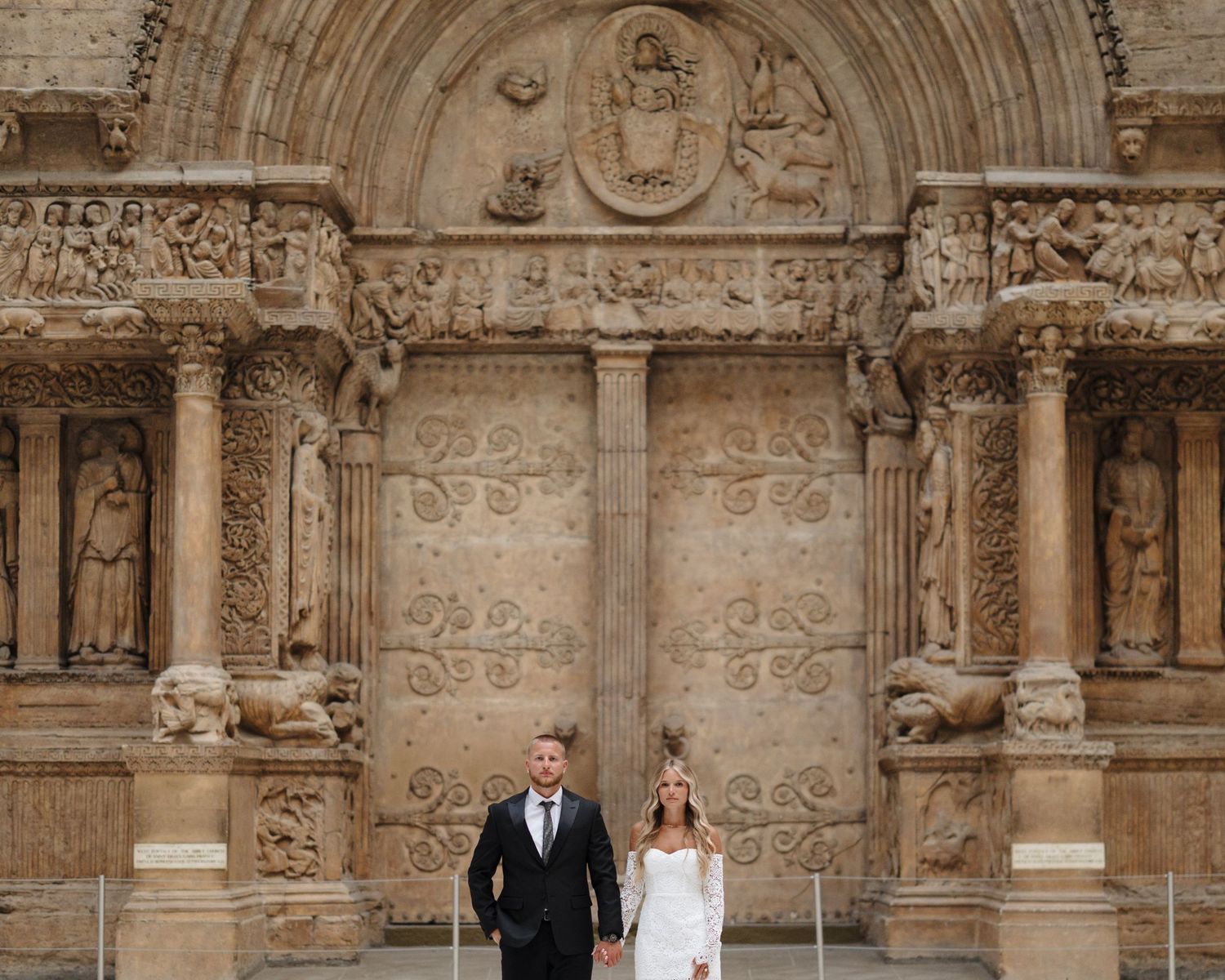 A couple in wedding attire stands at the entrance of an ornate stone cathedral with detailed archways and carvings.