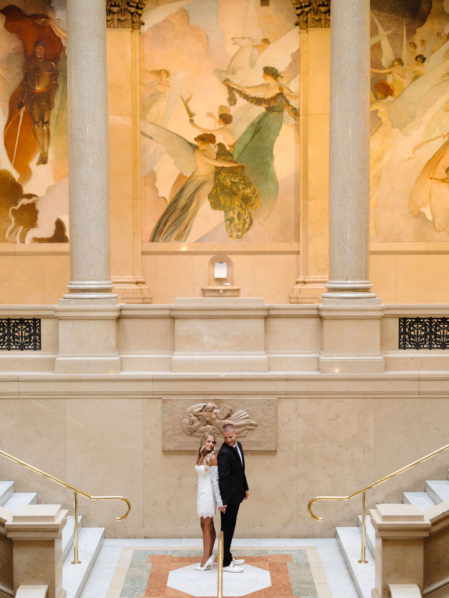 A couple shares a romantic moment on marble steps beneath ornate classical artwork and columns.