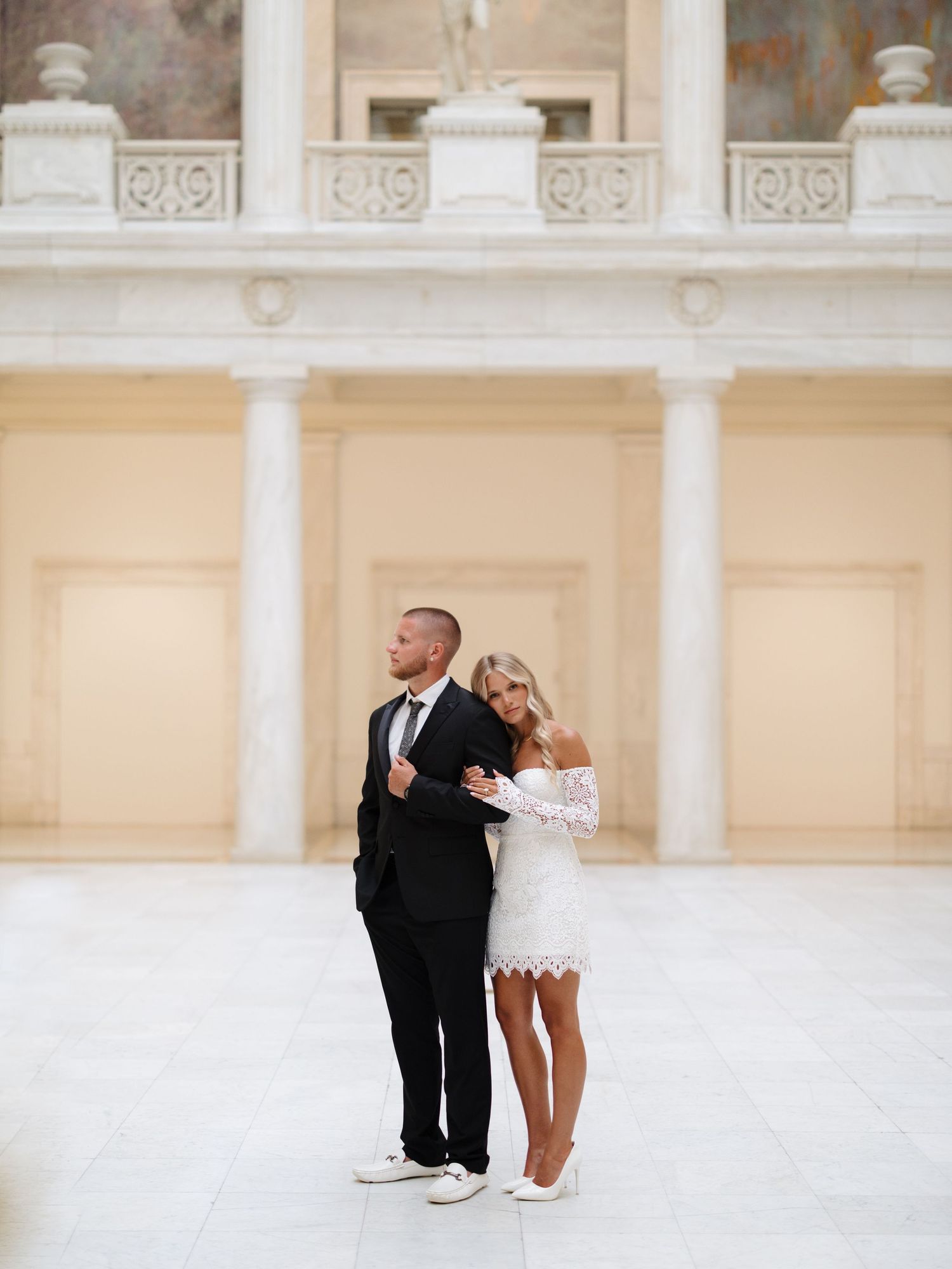 A couple in white and black formal attire pose together against elegant classical architecture with columns.
