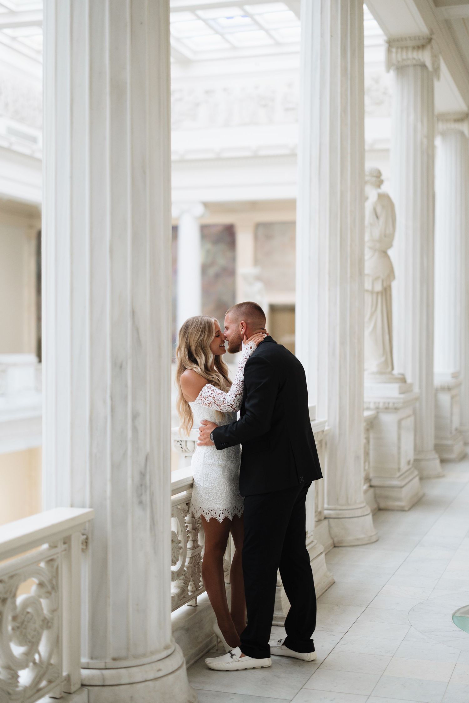 Romantic wedding couple shares intimate moments between elegant white columns in a bright colonnade.