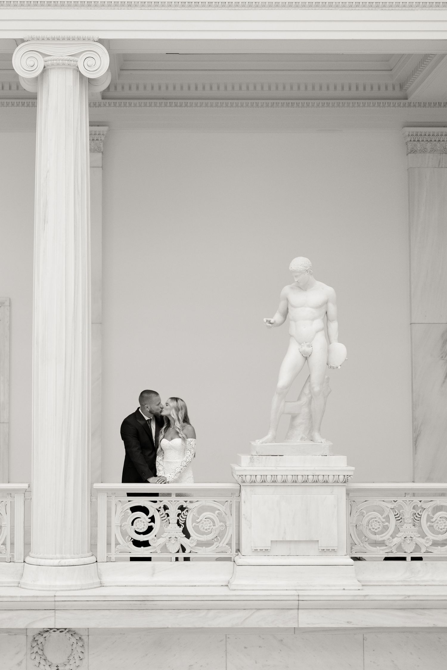 A classic black and white photo capturing a couple beside a white marble statue in a grand hall.