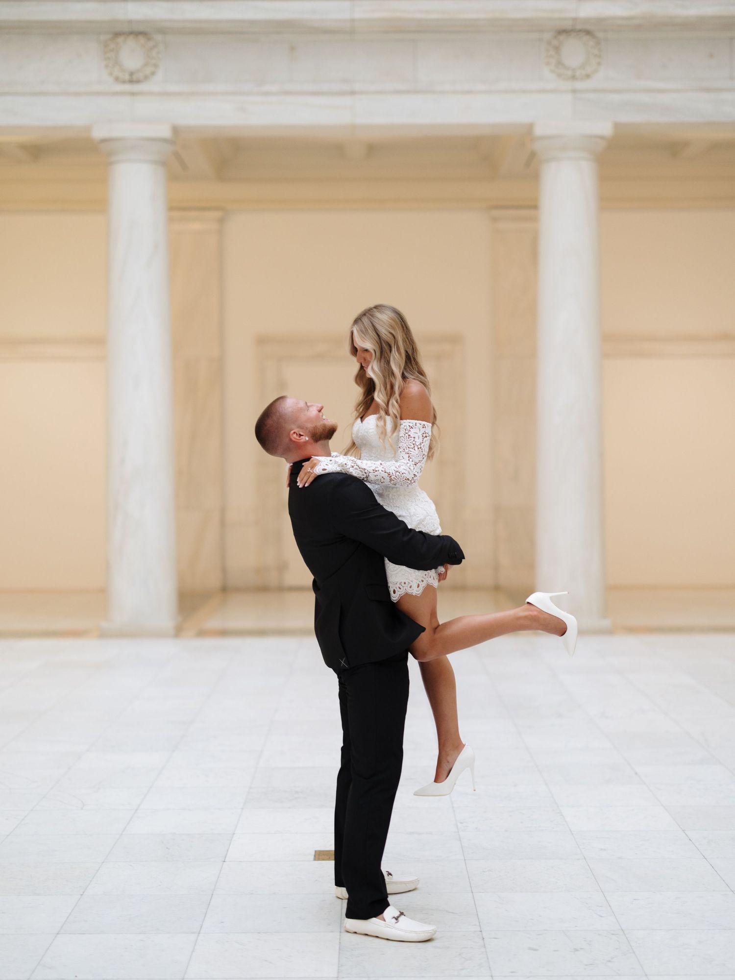 A romantic couple embraces in front of classic white columns, with the woman in white and man in black formal attire.