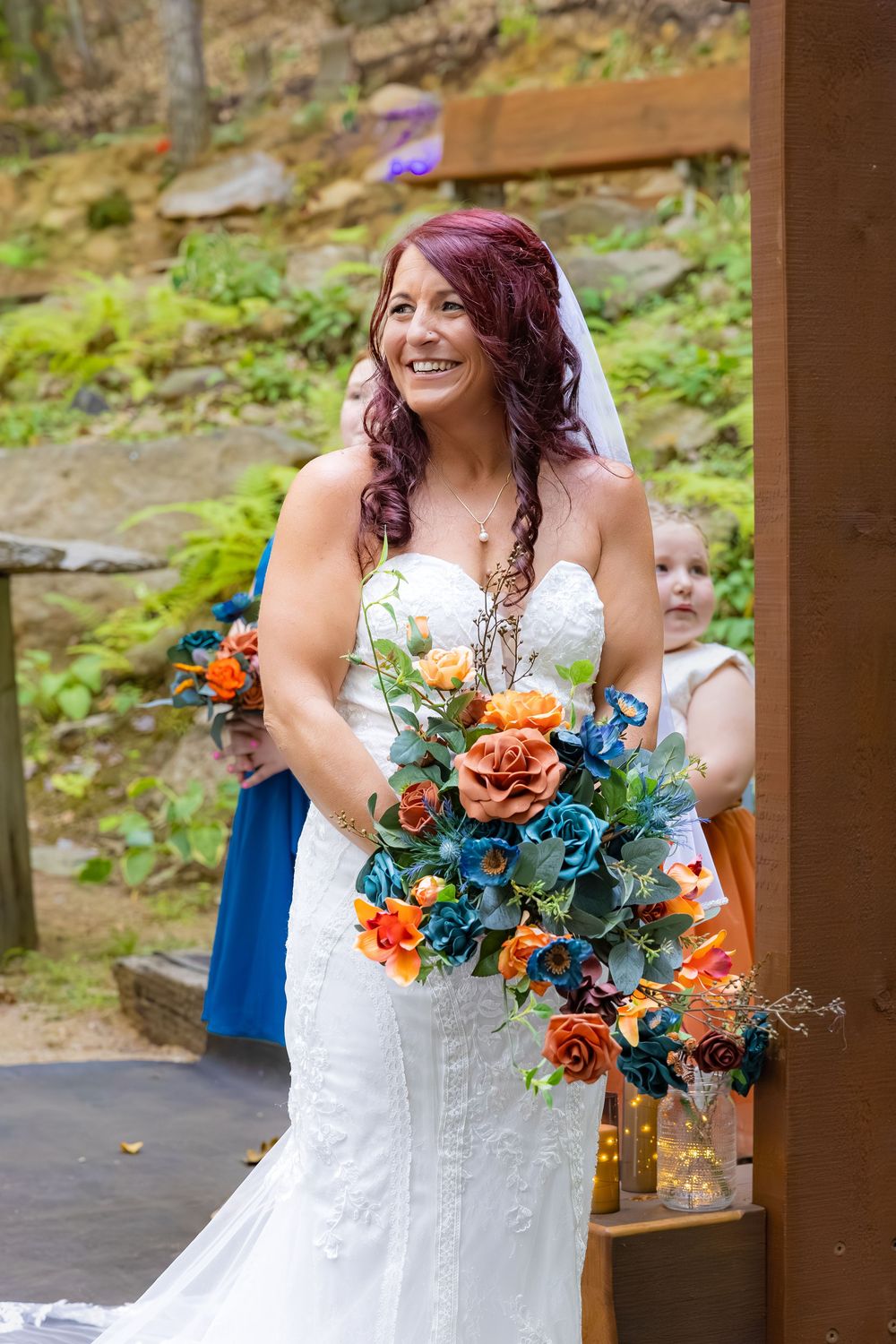 A bride in a white lace dress holds a vibrant orange and blue wedding bouquet while standing under a wooden gazebo.
