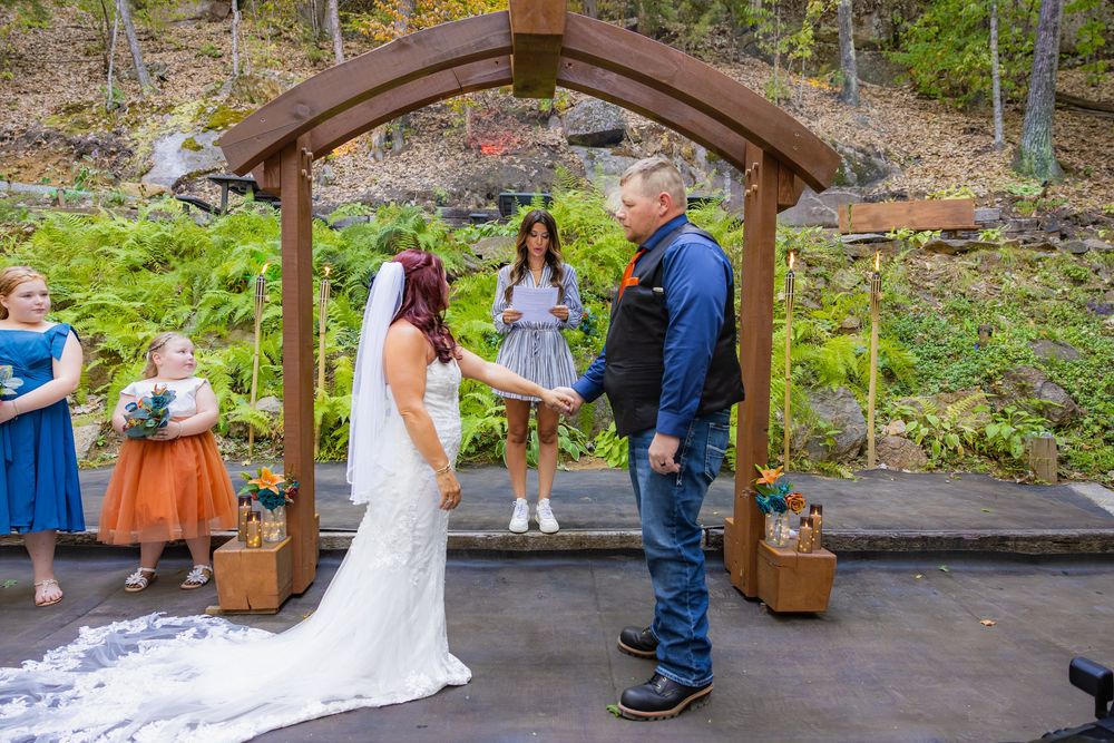 A couple exchanges vows under a rustic wooden arch surrounded by lush greenery and gardens during an outdoor ceremony.