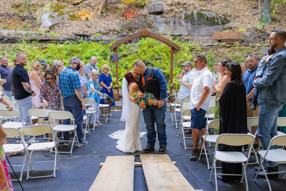 Wedding guests gather at outdoor reception venue with white folding chairs and tables arranged beneath tall trees.