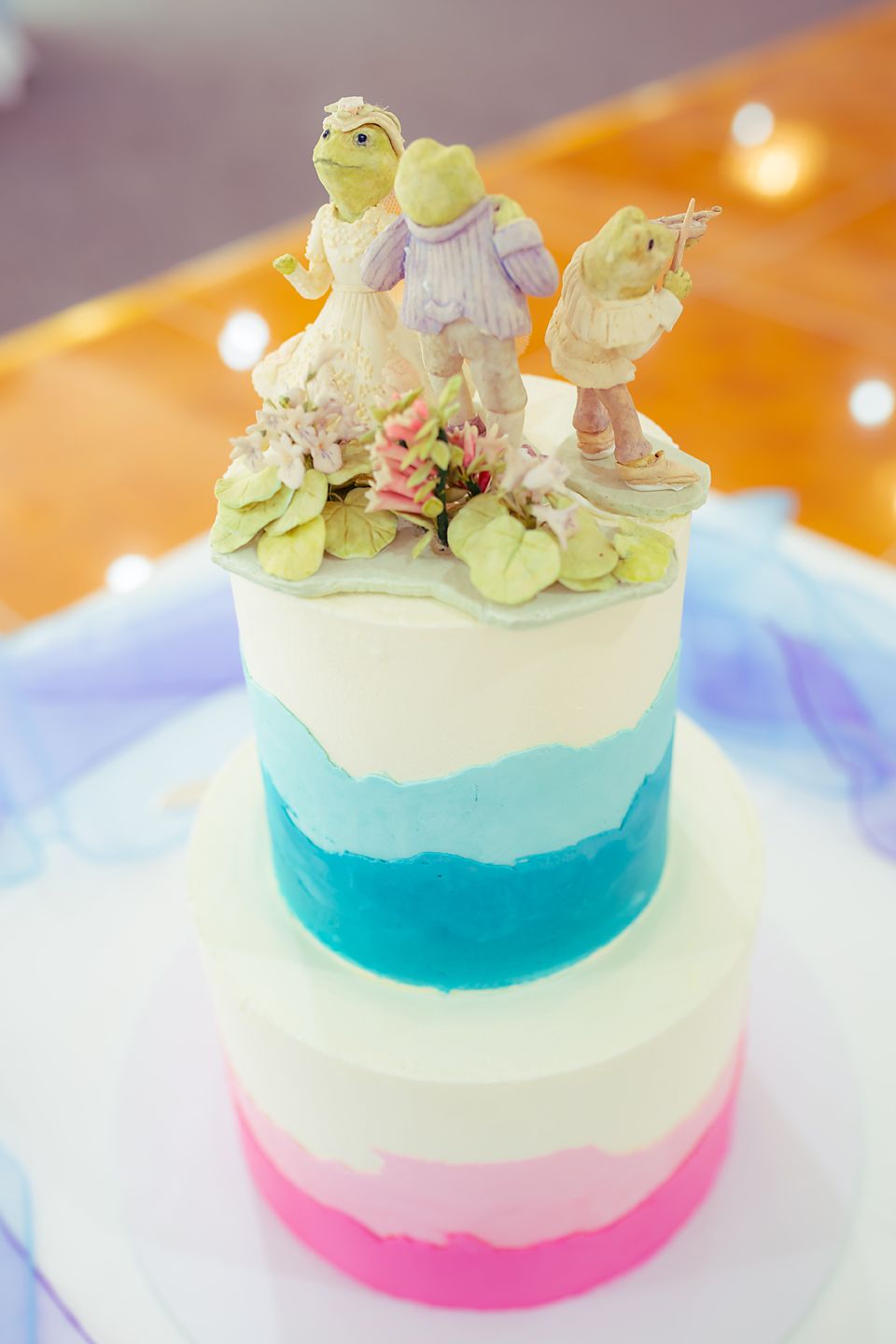A tiered wedding cake decorated in pink, blue and white ombre colors with whimsical figurine cake toppers on top.