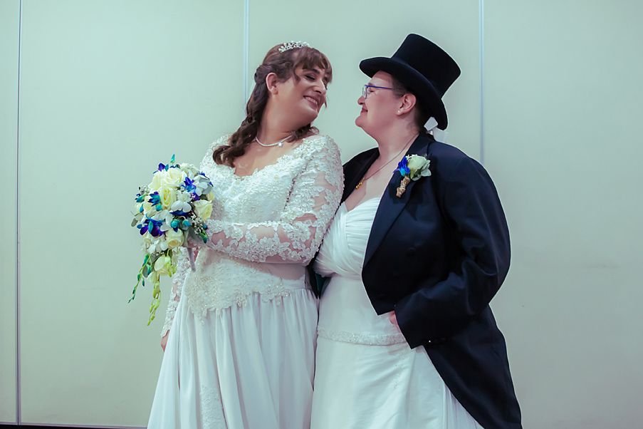 A couple in wedding attire shares a tender moment against a light green wall, one in white lace and one in black formal wear.