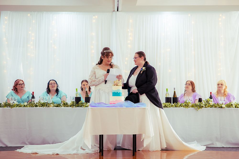 Wedding party sits at a long table during reception with white tulle and decor in background.