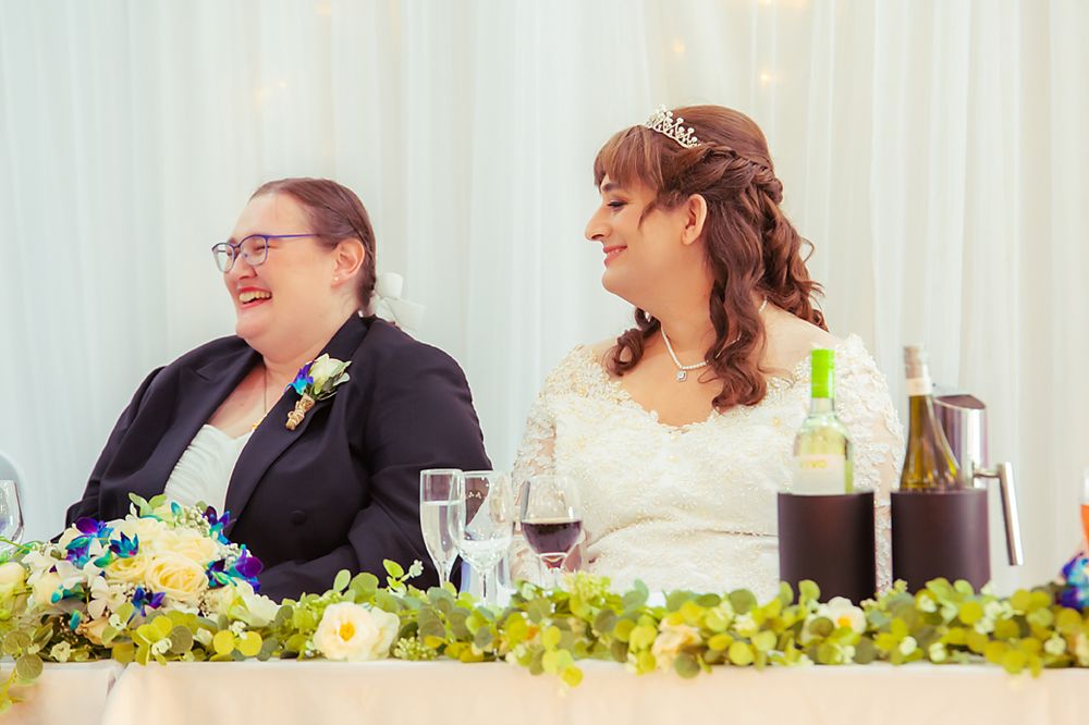 People seated at a wedding reception head table decorated with yellow and blue floral arrangements and white linens.