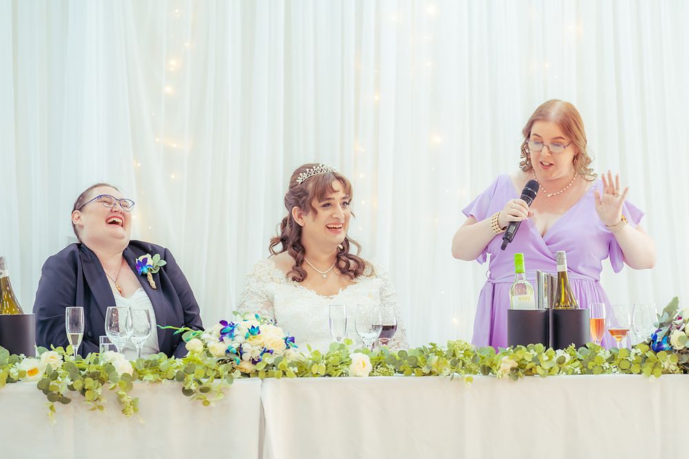 Long table at a wedding reception with guests celebrating with drinks and laughter among floral centerpieces.