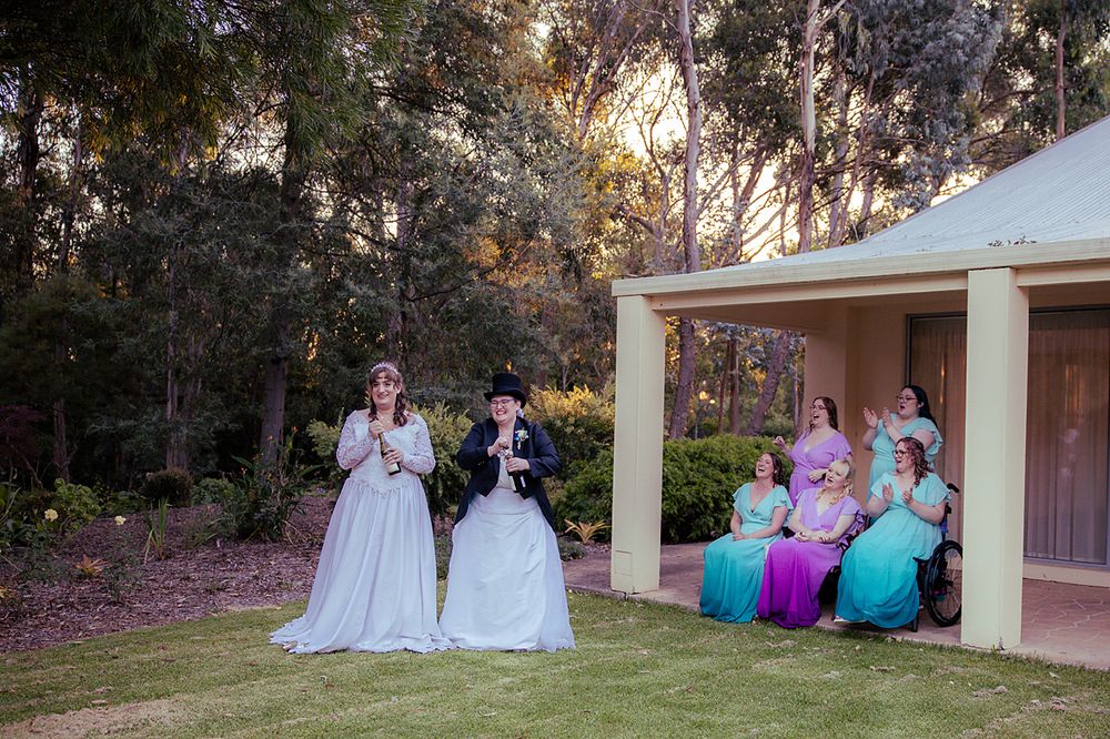Wedding party group sits together on lawn at sunset with elegant dresses and formal attire visible against outdoor backdrop.