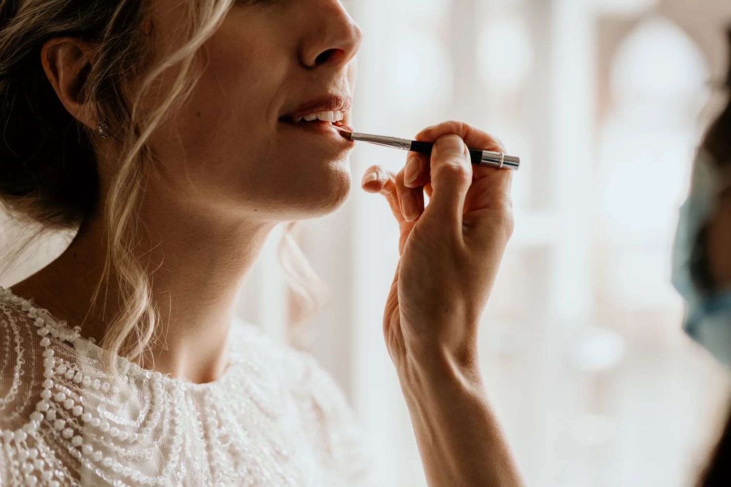 Close-up shot of makeup being applied with a brush to a bride's face against a light background.