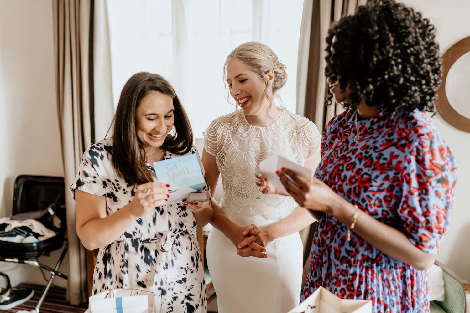 Three women share excited reactions while looking at something during wedding preparations.