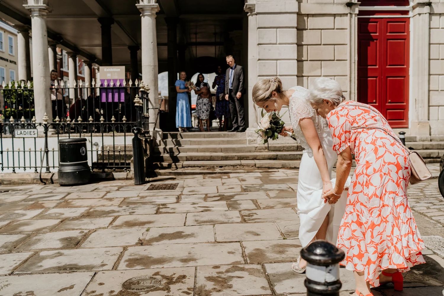 Two people in elegant attire bend down to pet a dog on stone steps outside a historic building.