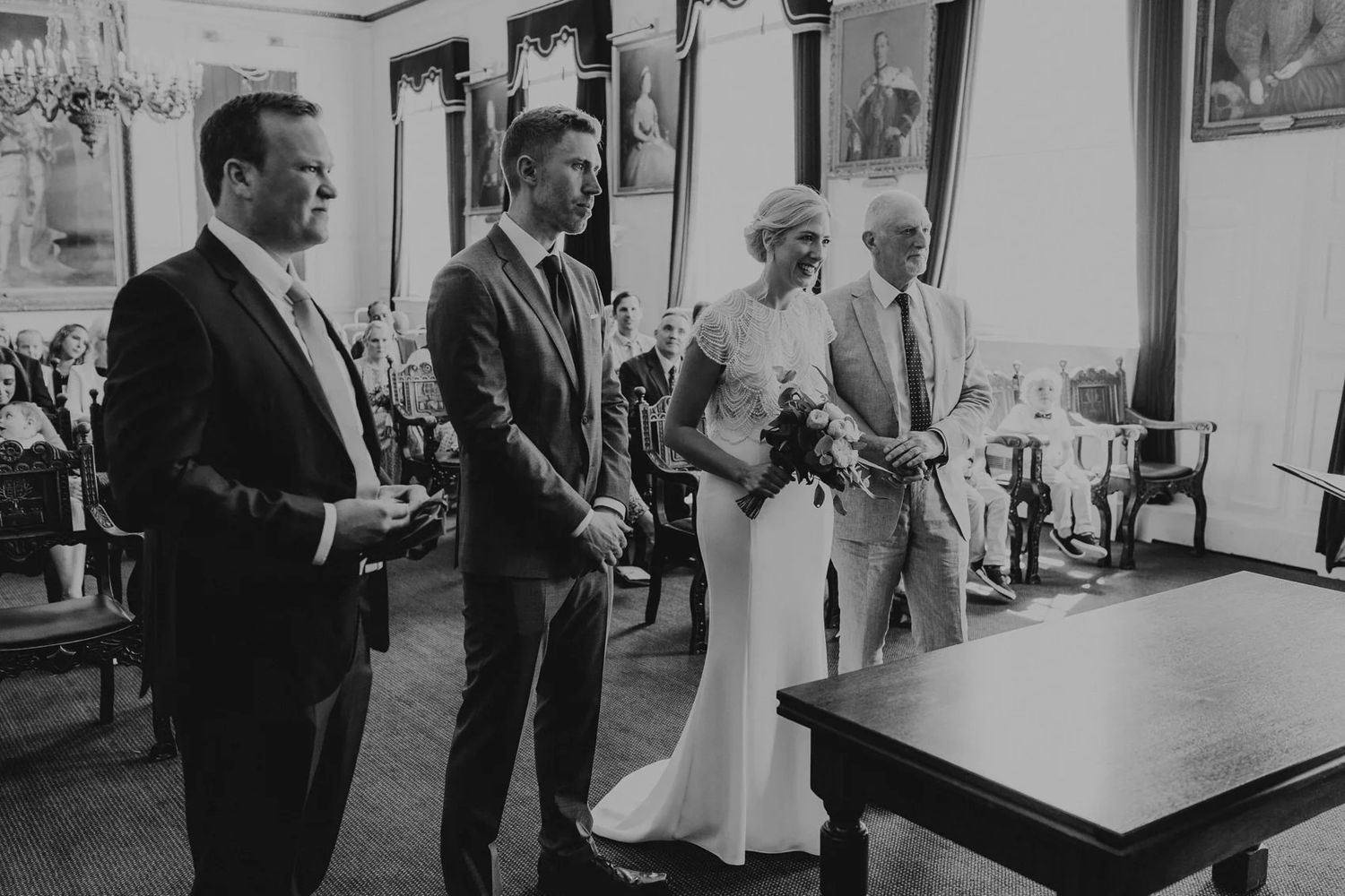 A black and white wedding photograph showing a group standing in formal attire indoors.