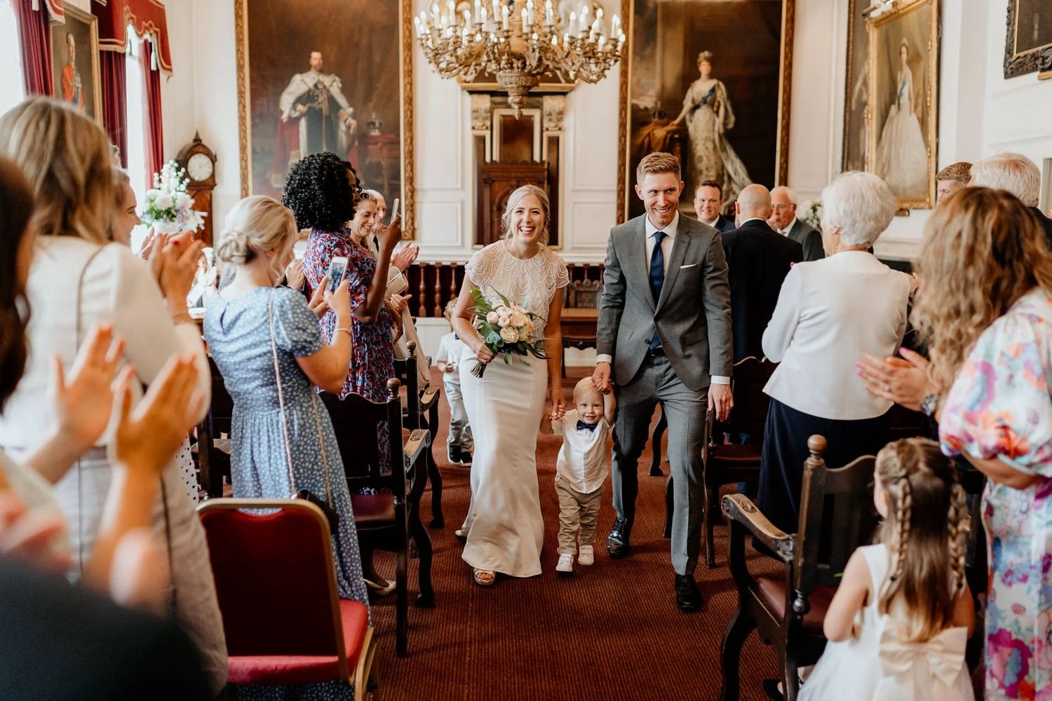 Wedding guests celebrate and dance in a grand historic room with crystal chandeliers and classic decor.