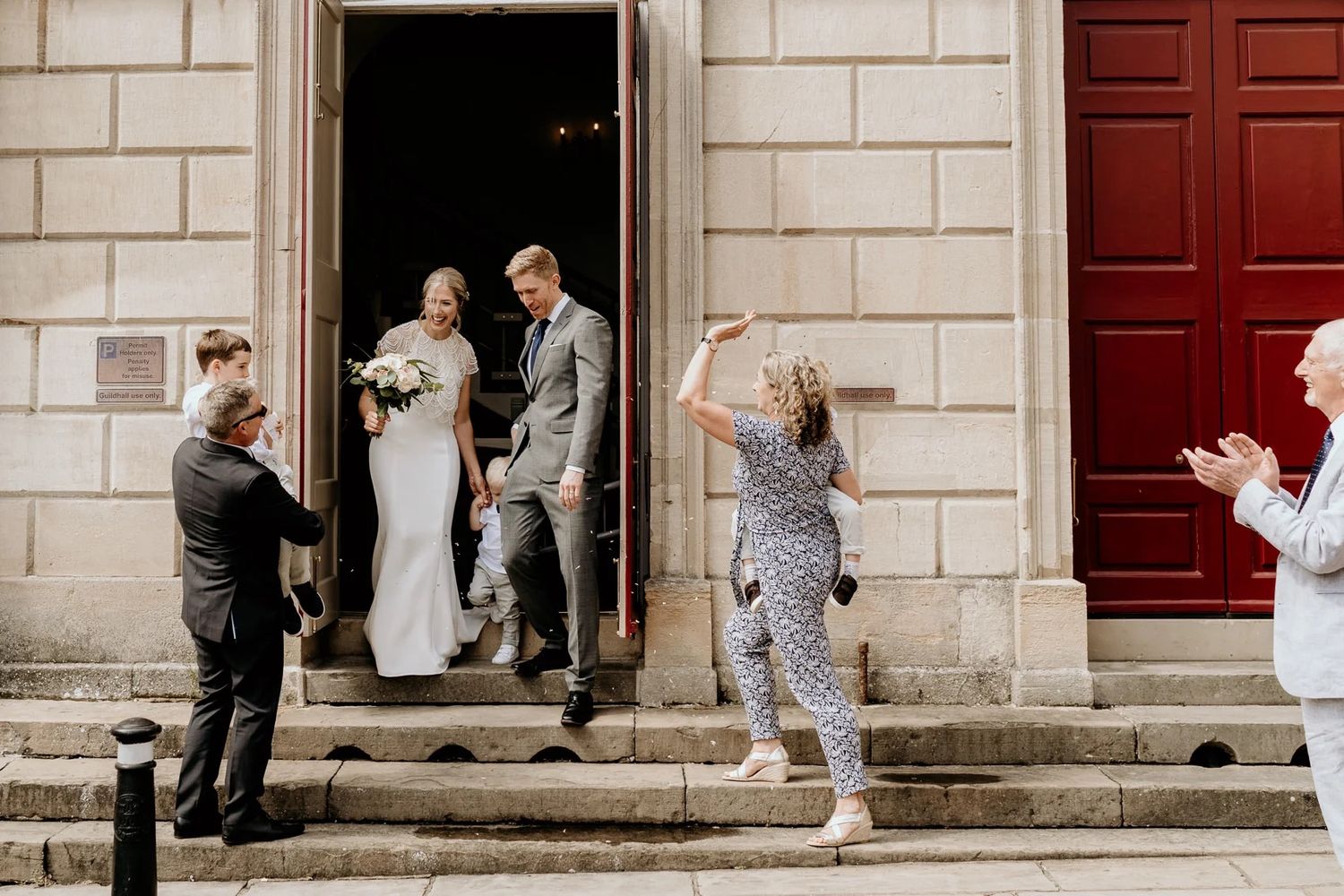 Wedding party members gather on stone steps outside a building with red doors.