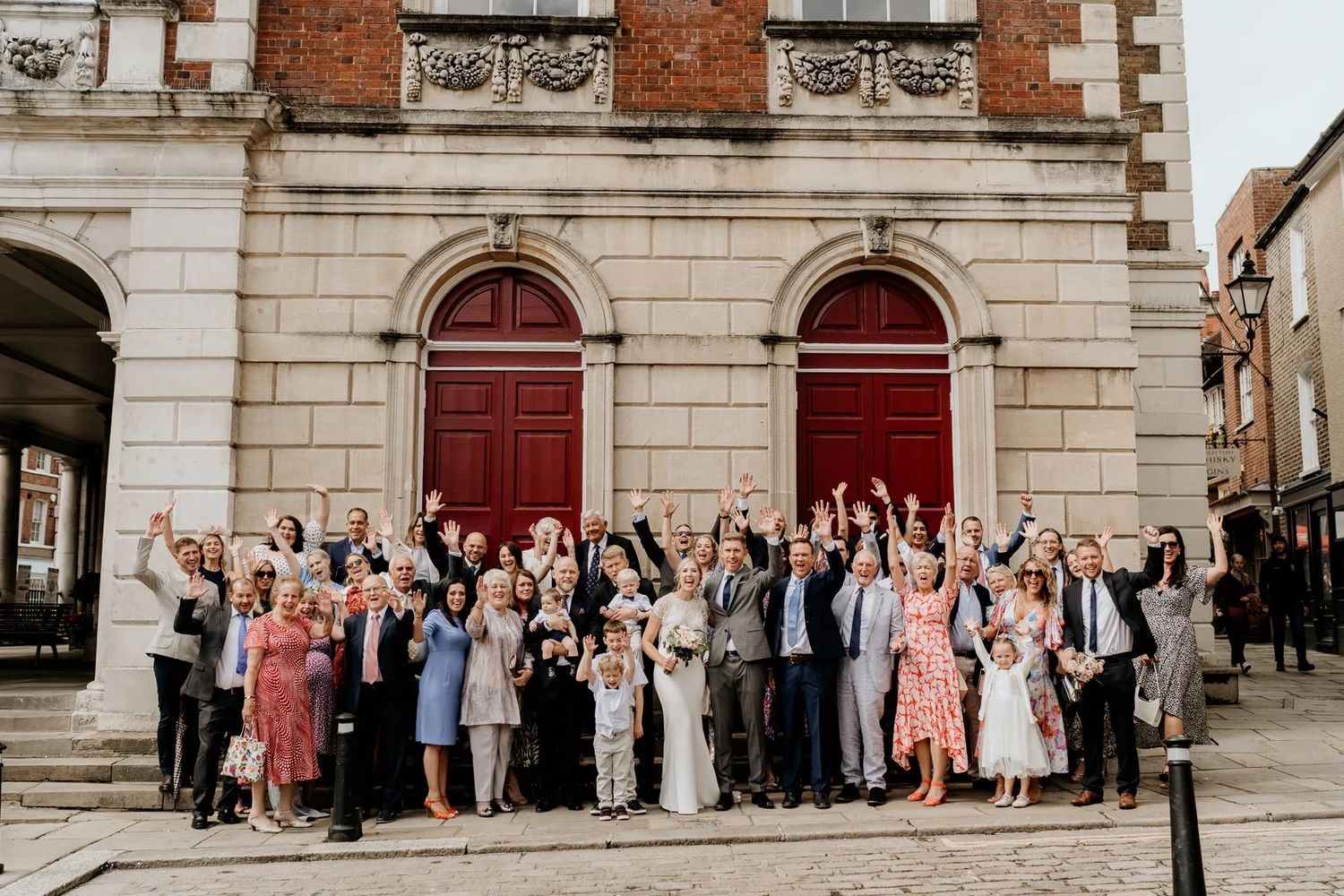 Large group photo of wedding guests outside a classic stone building with red doors.