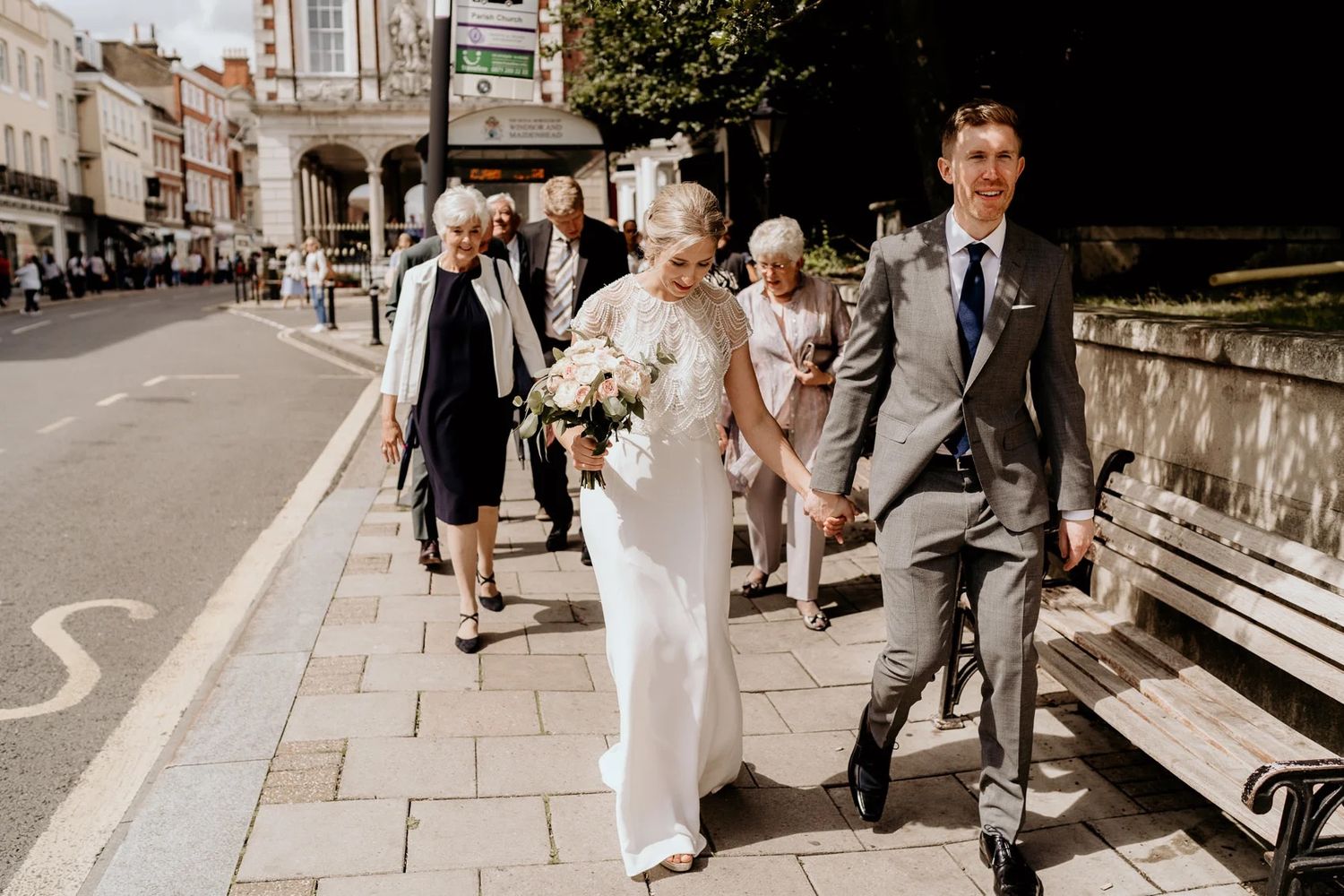 Wedding party walking along historic stone buildings on sunny day.