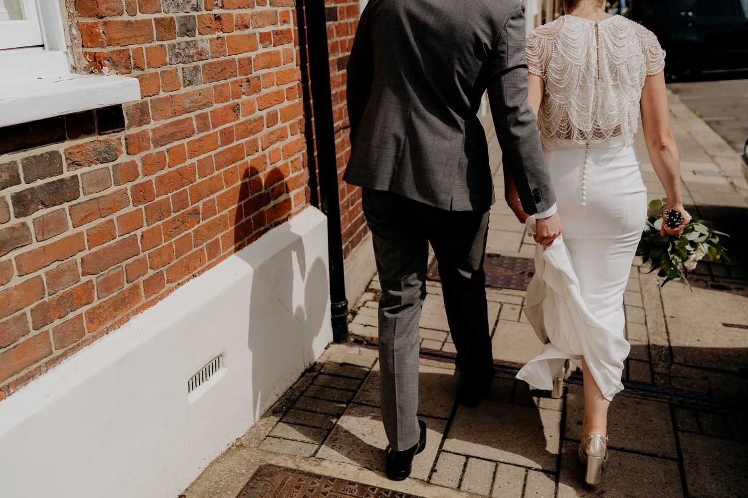 Bride and groom walking together along a brick wall exterior in formal wedding attire.