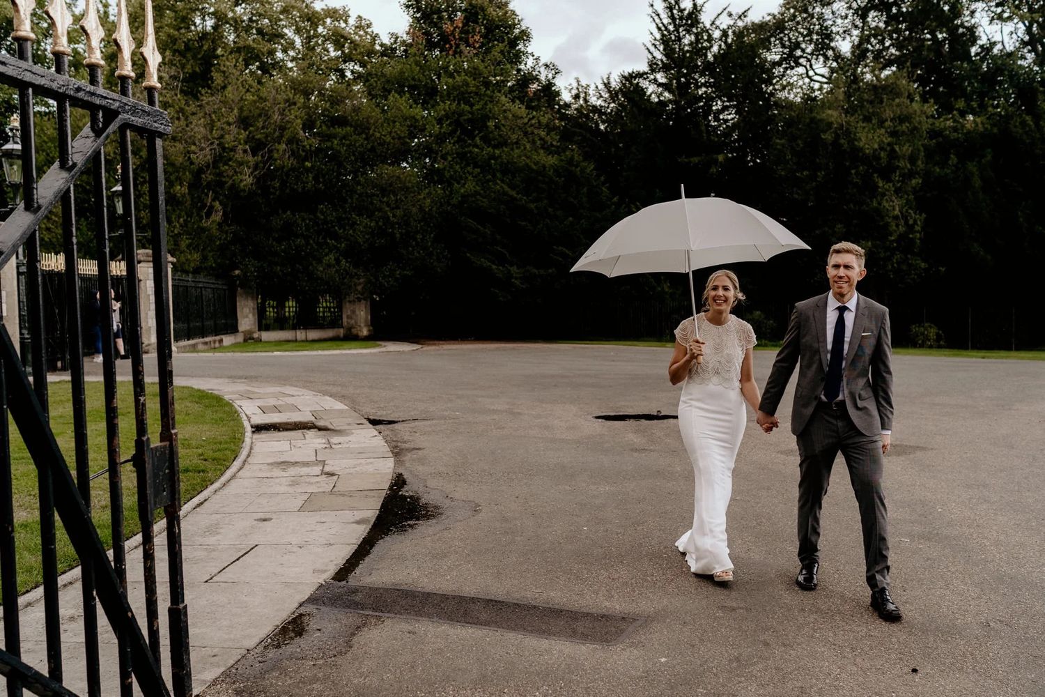 Two people walk down a path with a white umbrella in an outdoor venue during a celebration.