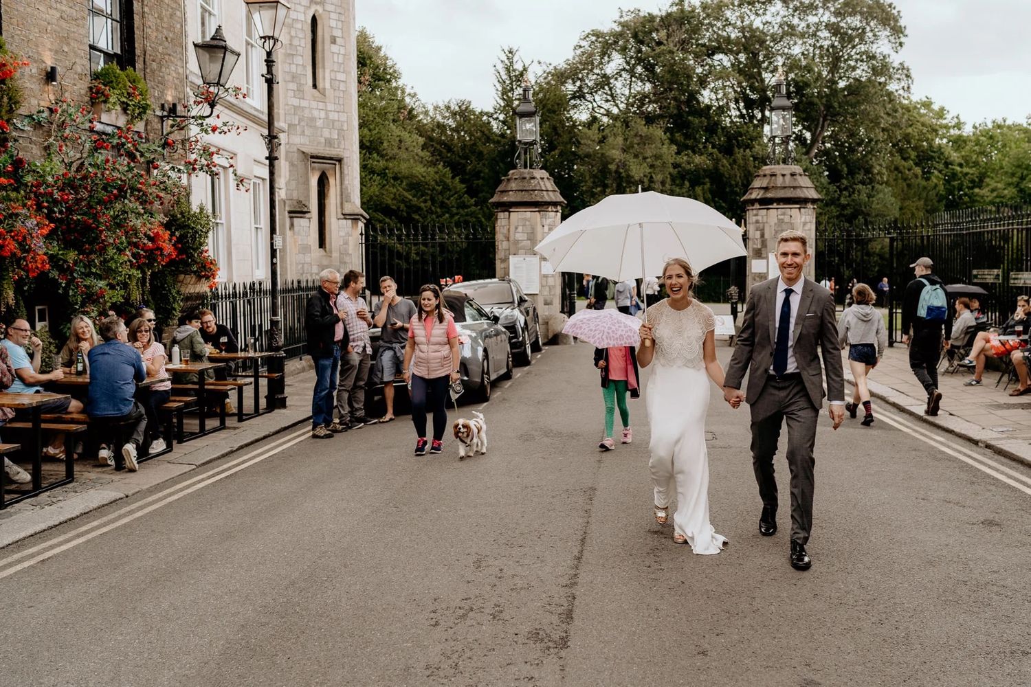 A wedding group walks down an old cobblestone street lined with historic buildings and lamp posts on a sunny afternoon.