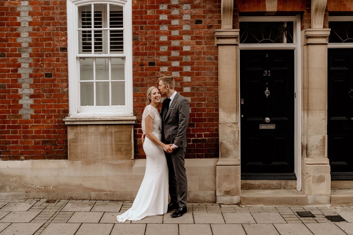 Romantic wedding couple poses in front of historic brick building with white windows and stone accents.