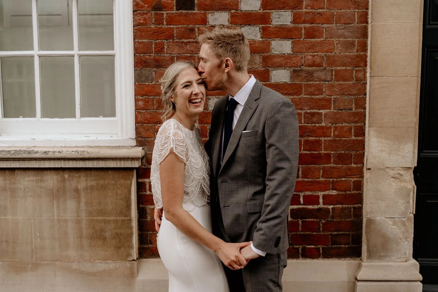 Happy couple shares a joyful moment during wedding photos against rustic brick wall backdrop.