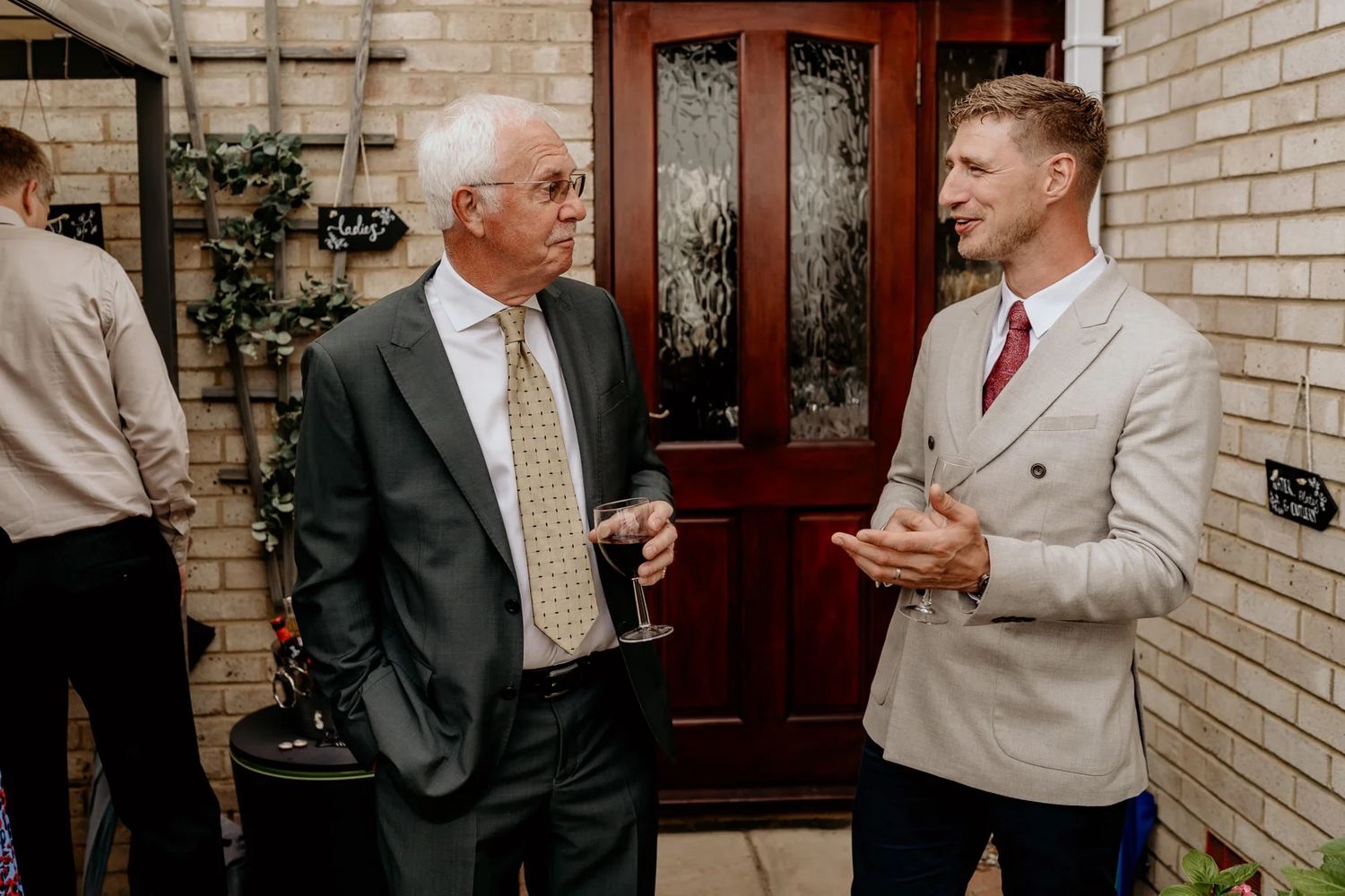 Wedding guests in formal attire chatting and socializing outside a brick building.