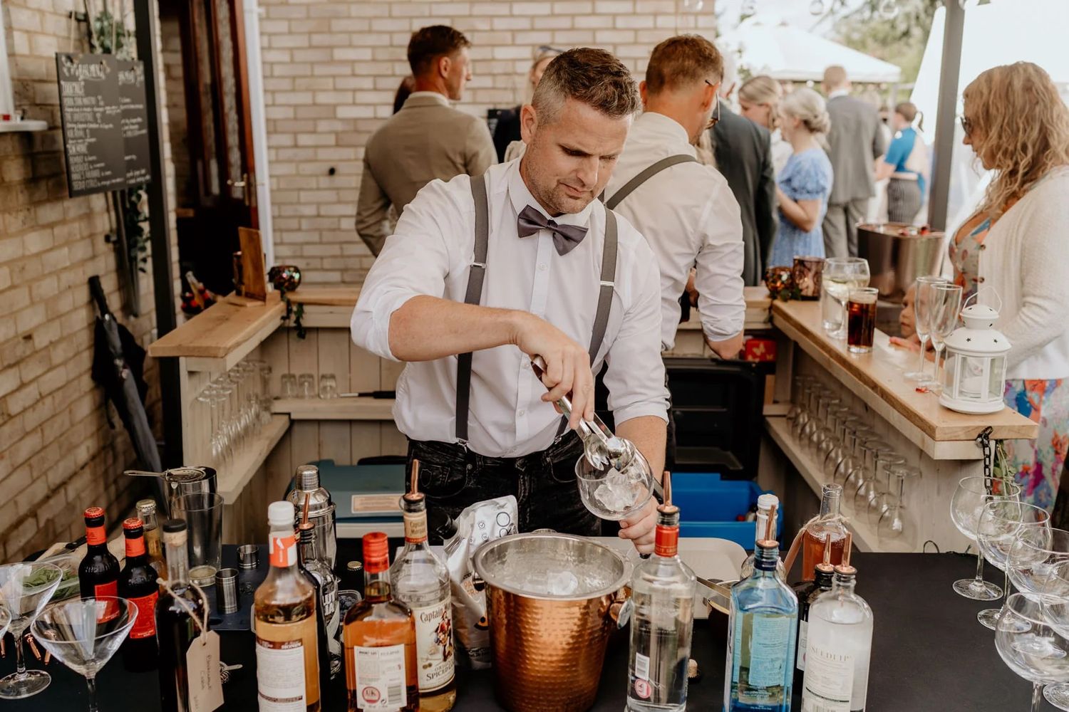 Wedding guests gather at a rustic brick-walled bar for cocktails and refreshments during an elegant reception.