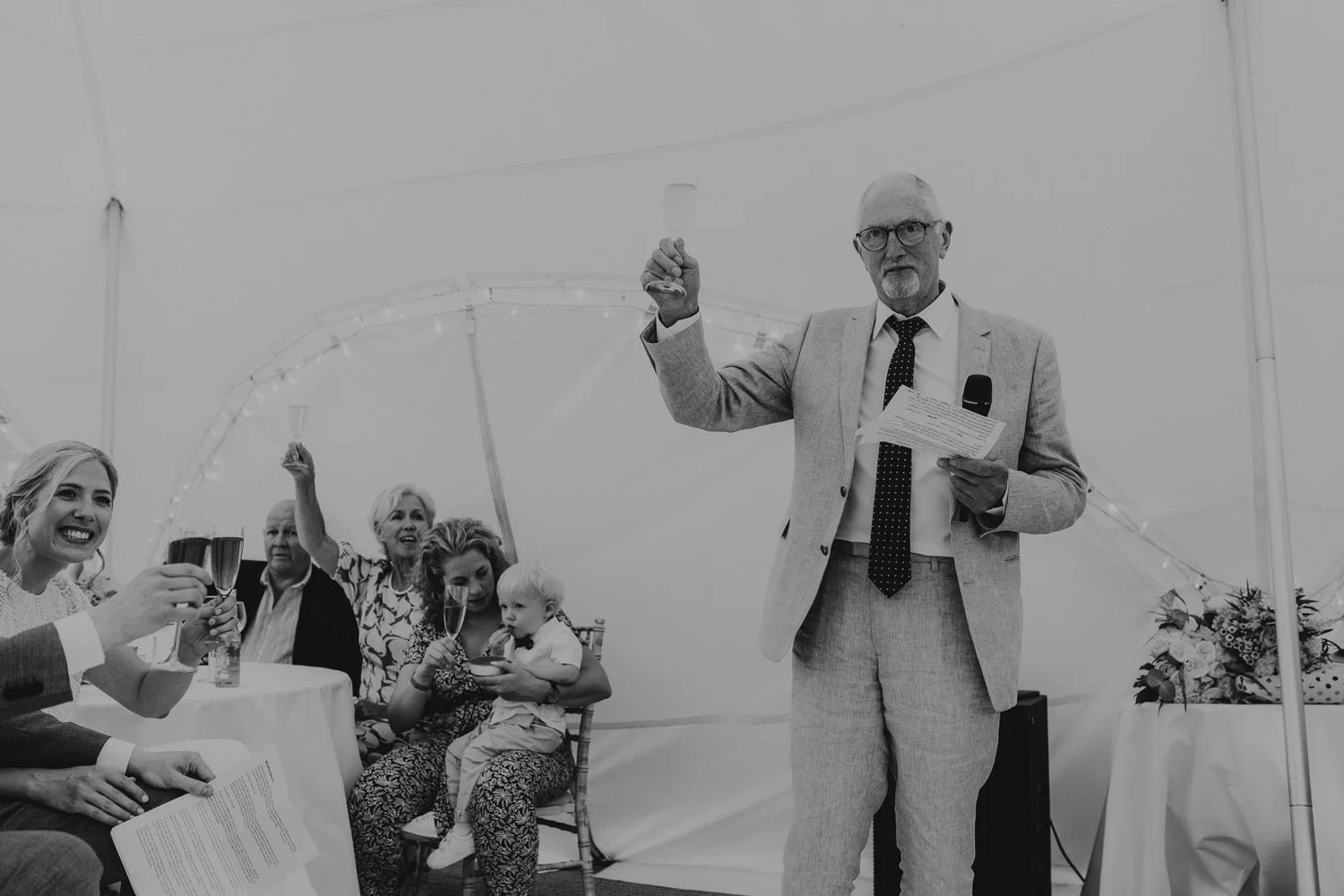 Black and white photo of wedding guests mingling under a white tent during an outdoor celebration.