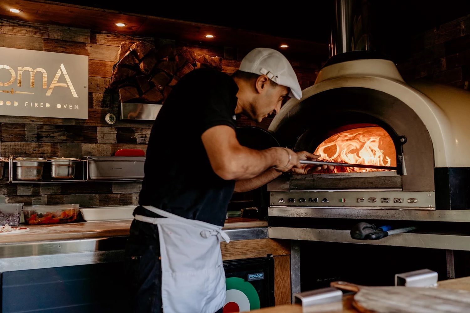 Professional chefs working in a dimly lit restaurant kitchen with pizza ovens.