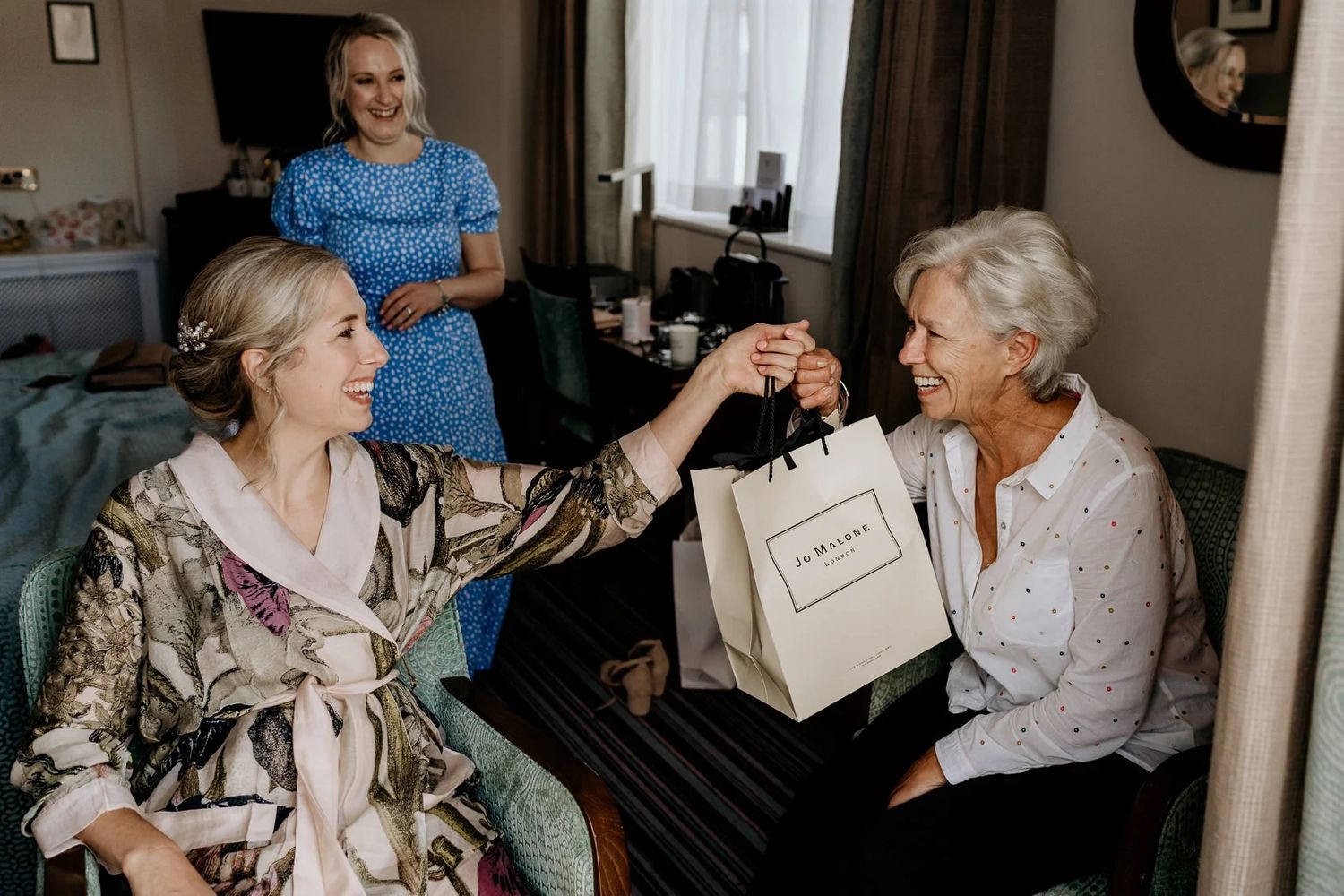 Three generations of women share a joyful moment while exchanging a gift bag in a living room.