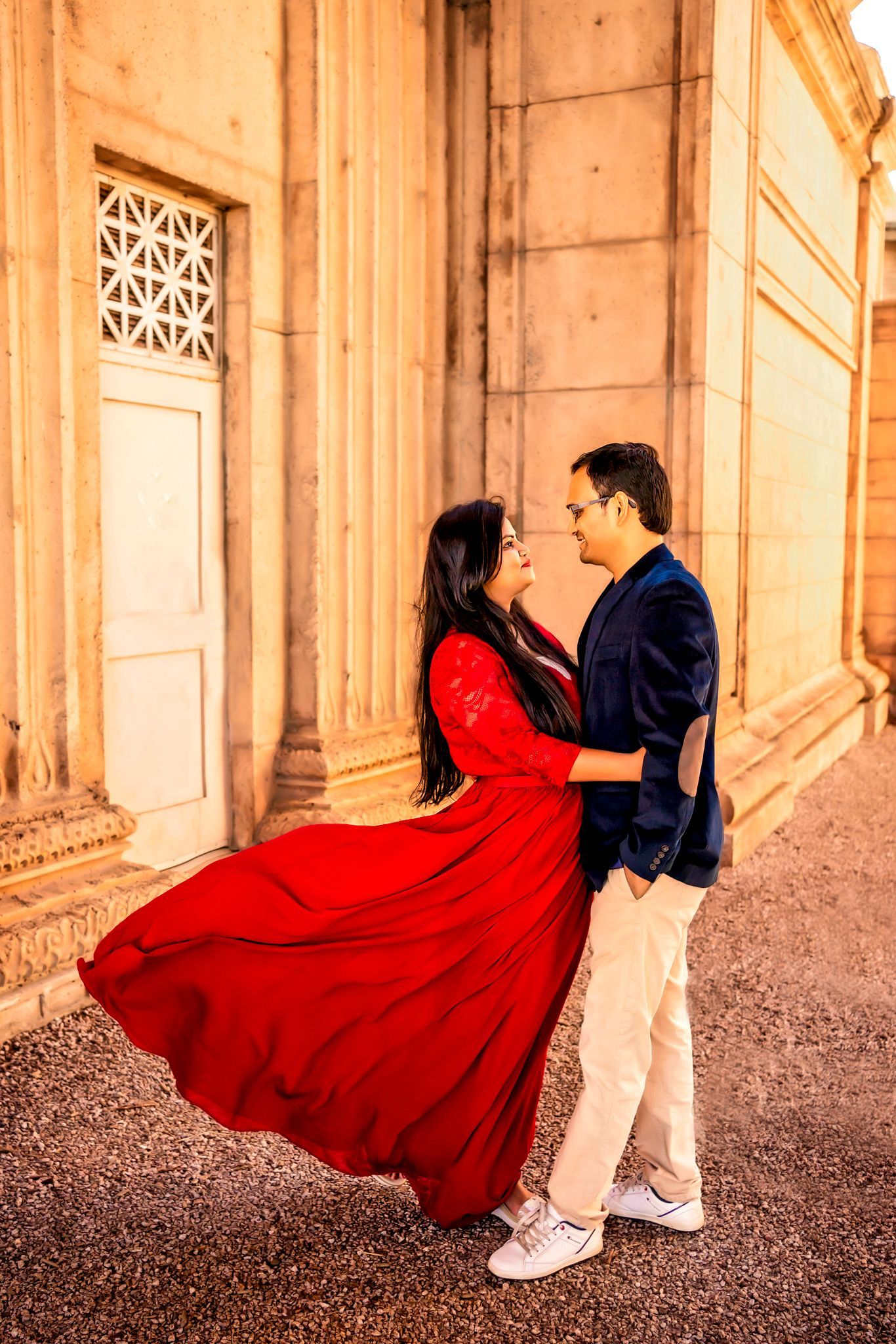 A couple in red dress and navy suit dance in a romantic sequence against a rustic architectural backdrop.
