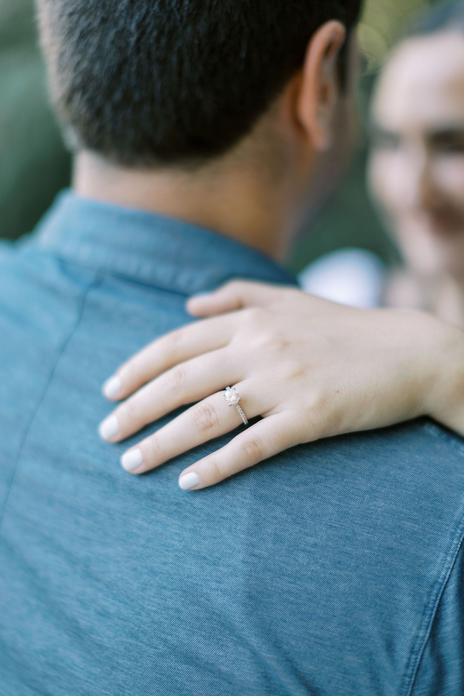 A woman's hand with an engagement ring rests on a person's shoulder wearing a blue shirt.